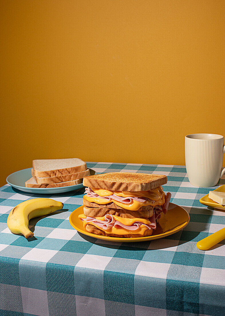 A homemade breakfast on a checkered blue tablecloth features a sandwich, banana, coffee, and butter against yellow background