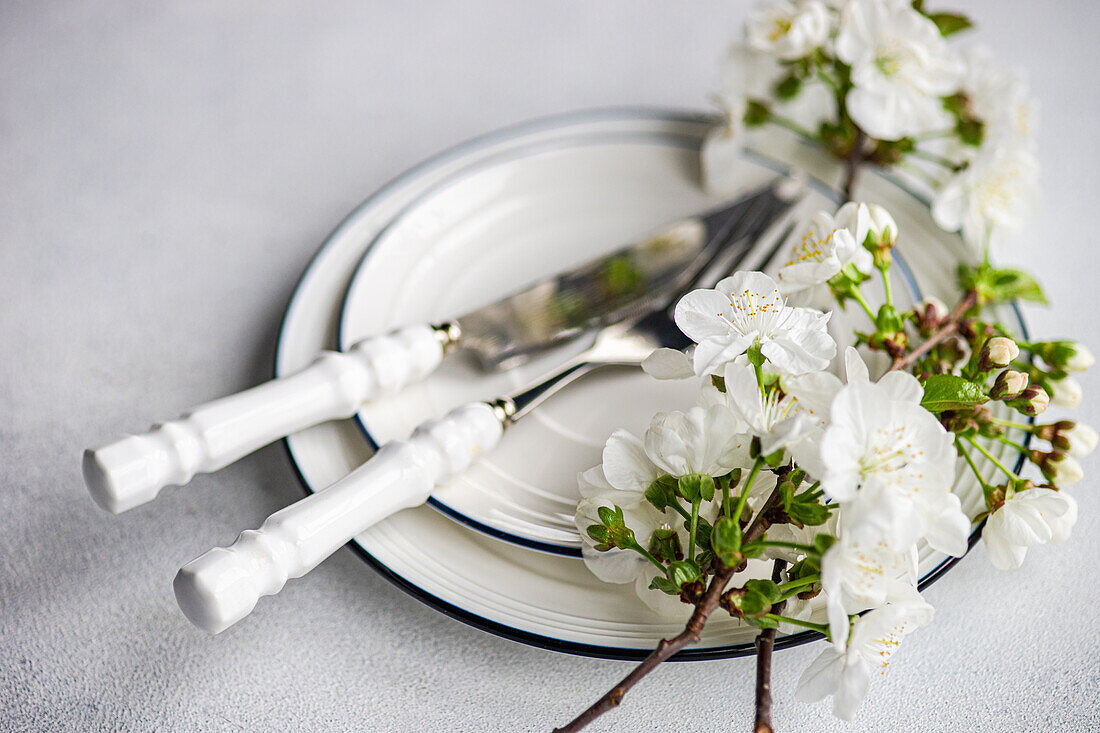 Eleganter Frühlingstisch mit weißen Kirschblüten auf einem Zweig, gepaart mit sauberen weiß und blau gestreiften Tellern und Silberbesteck mit weißen Griffen, die eine frische, saisonale Atmosphäre schaffen