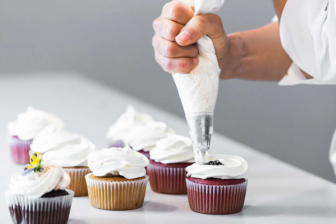 Ein nicht erkennbarer Bäcker drückt Schlagsahne aus einem Spritzbeutel auf leckere Cupcakes, während er in der Backstube Desserts zubereitet