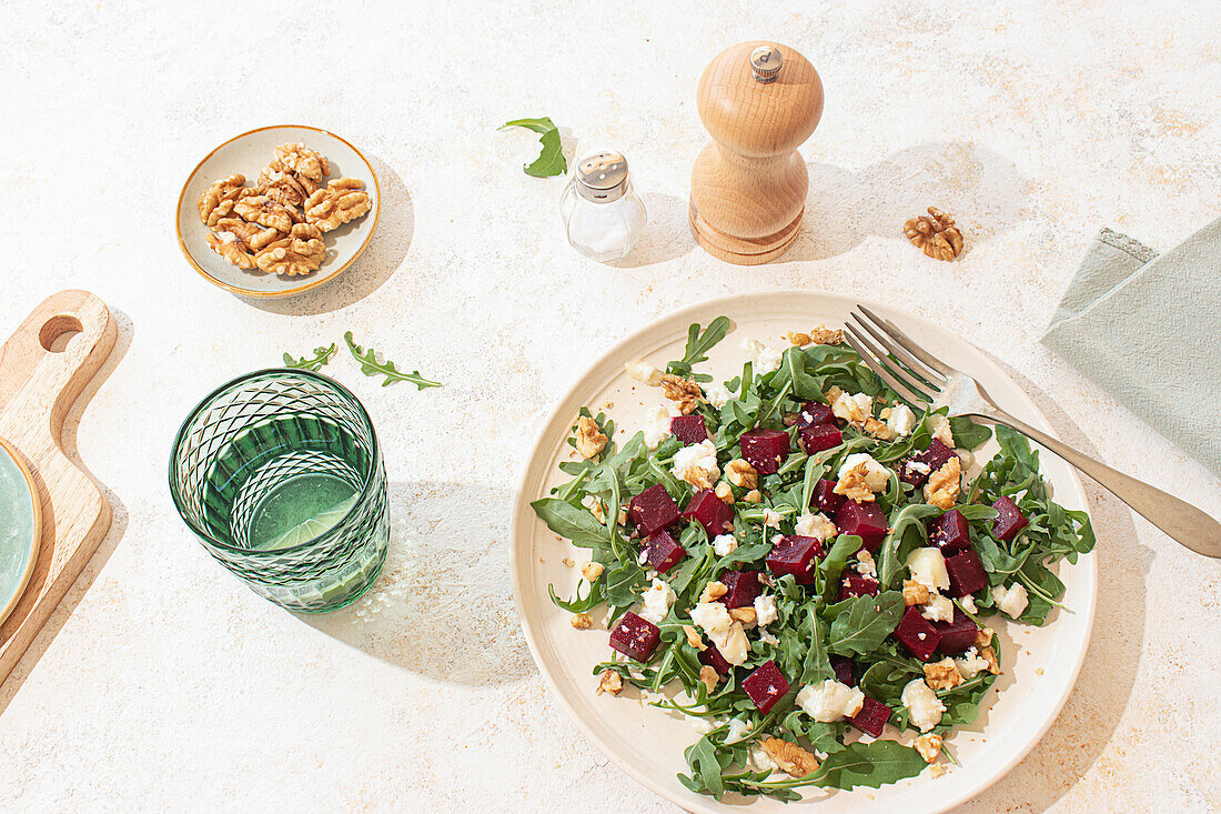 Gesunder Salat aus Ziegenkäse, Rucola und Roter Bete auf einem beigen Teller mit einer Pfeffermühle und einem Kalkwasserglas in rustikalem, aber lebhaftem Ambiente