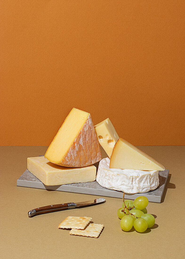 From above, a variety of cheeses including Brie, Swiss, and Cheddar, paired elegantly with crackers and grapes, positioned on a textured beige surface alongside a rustic cheese knife