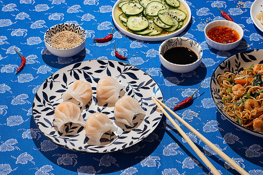 Vielfältige chinesische Küche mit Teigtaschen, Gurken in Essig, scharfen Soßen und gebratenen Nudeln, attraktiv angerichtet auf einer blauen, blumengemusterten Tischdecke in hellem Sonnenlicht