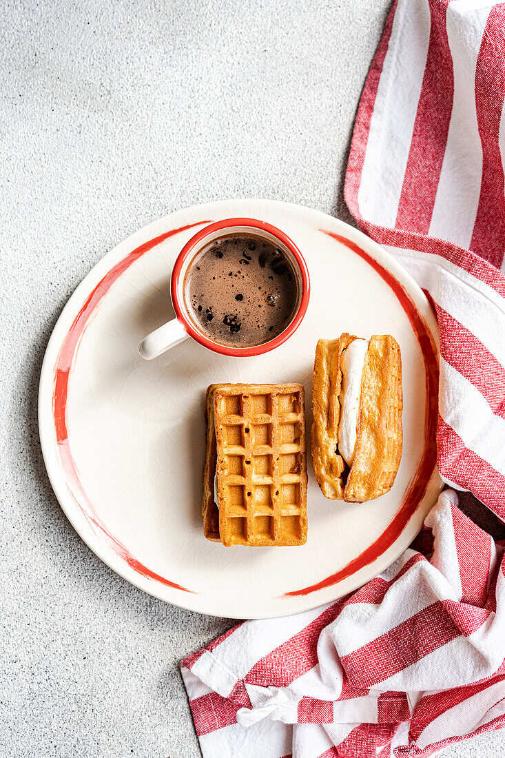 A top view of golden homemade waffles with a vanilla marshmallow filling alongside a cup of hot coffee on a rustic plate with a striped towel