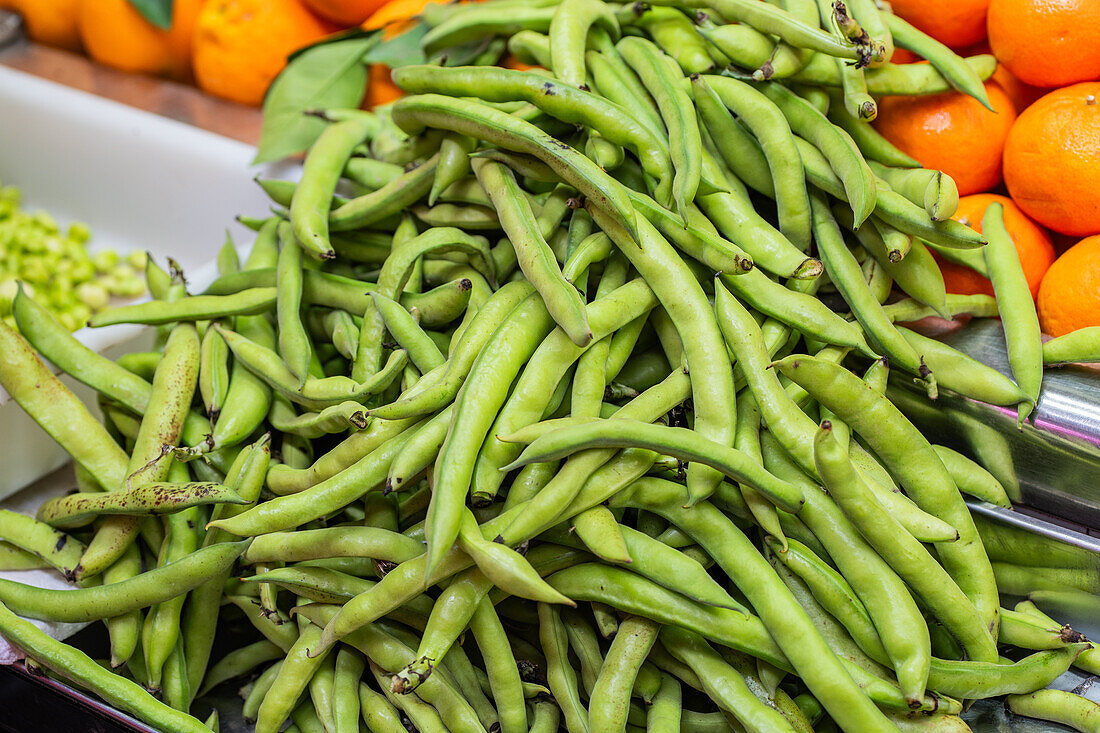 Frische grüne Bohnen, die auf einem Markt aufgestapelt werden, mit einem Hintergrund aus leuchtenden Orangen