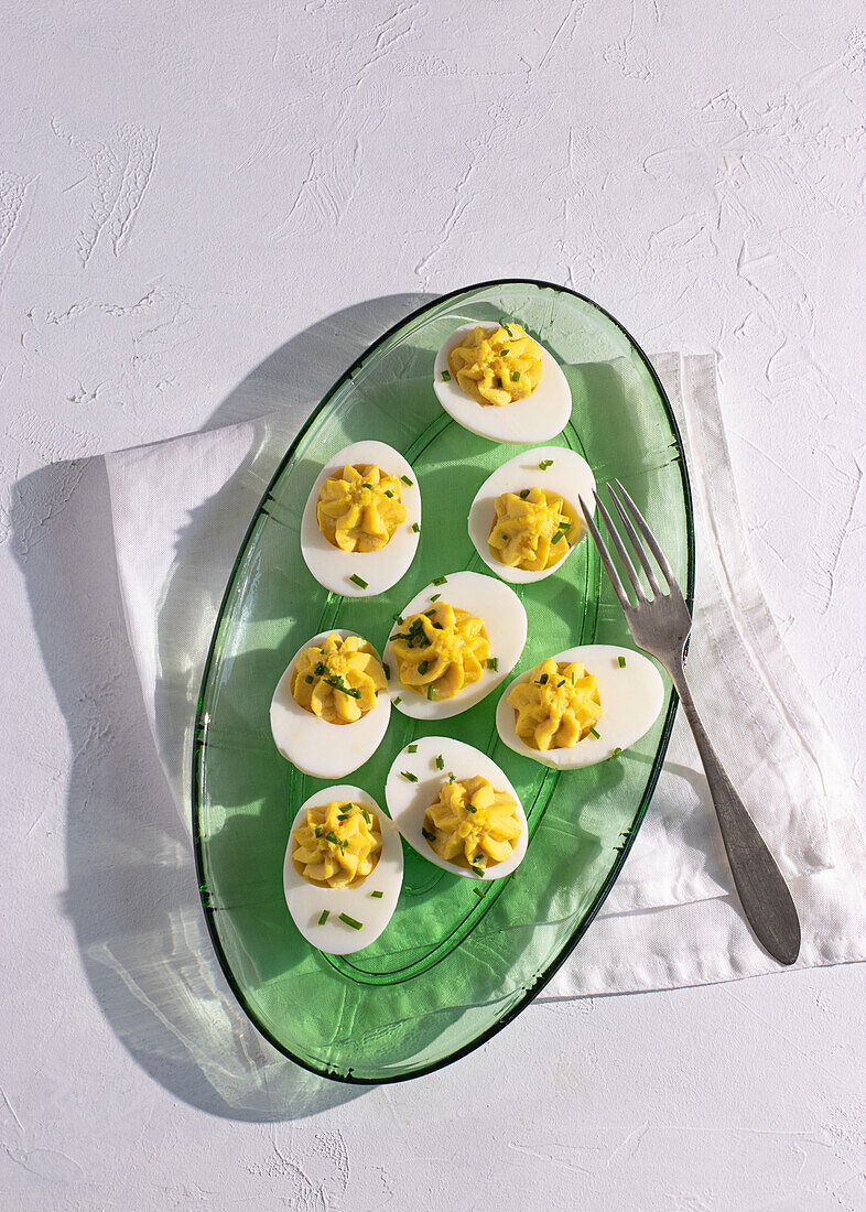 Draufsicht auf klassische "deviled eggs", serviert in einer transparenten grünen Glasschale, hervorgehoben durch natürliches Sonnenlicht, das weiche Schatten wirft, perfekt für eine Gourmet-Vorspeisenpräsentation.