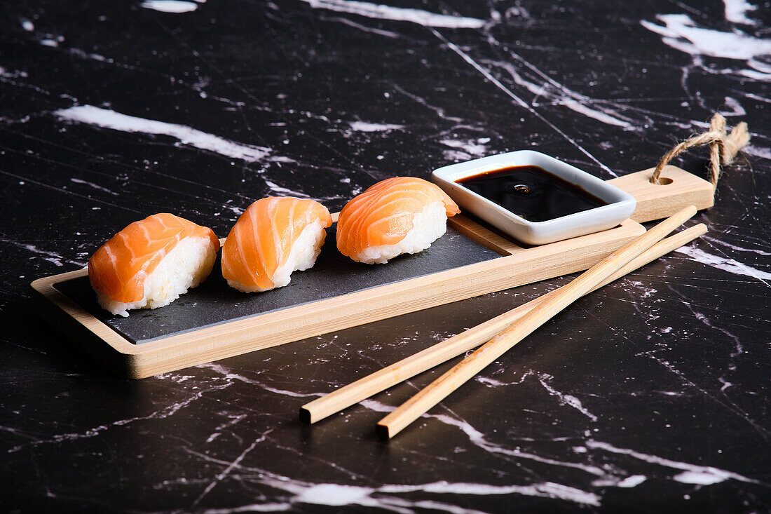 Three pieces of salmon sushi served on a wooden board with soy sauce and chopsticks on a marbled background.