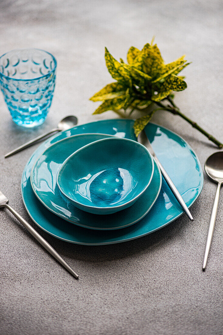 Stylish table setting featuring bright turquoise ceramic dishware and sleek cutlery on a textured gray surface. A decorative plant accompanies the scene, adding a touch of nature.