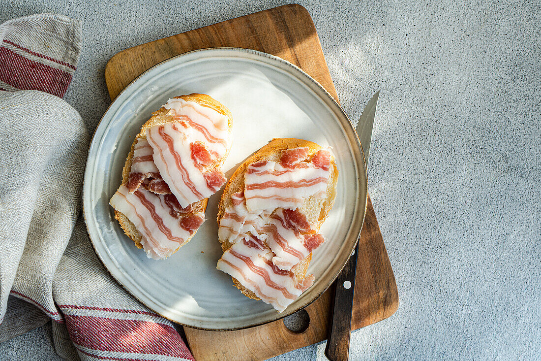 From above tow toasted bread topped with thin slices of lard, presented on a ceramic plate beside a wooden cutting board and knife.