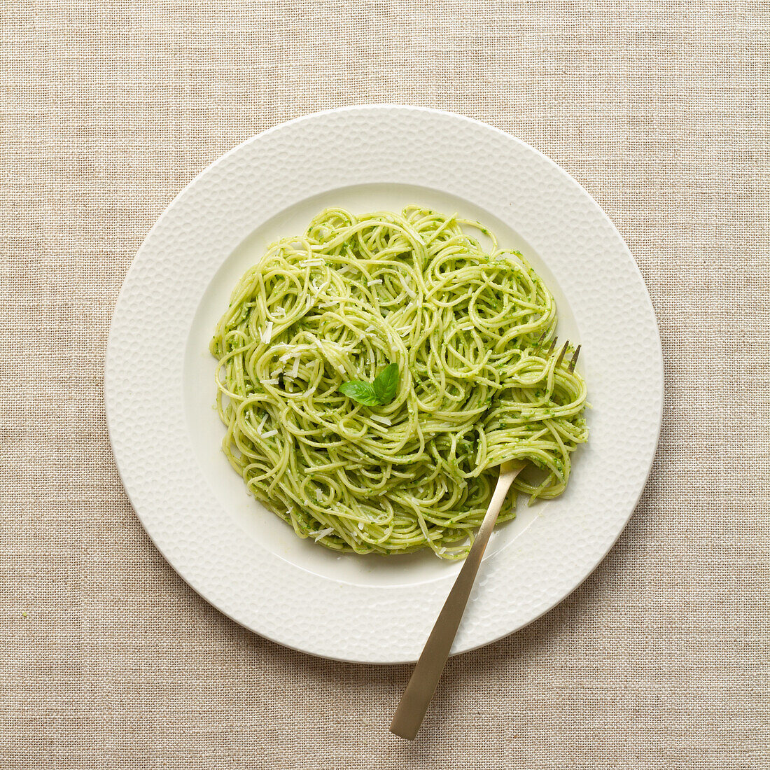 Draufsicht auf einen weißen Teller mit Spaghetti in leuchtend grüner Pestosauce, serviert auf einem beigen Leinentischtuch.