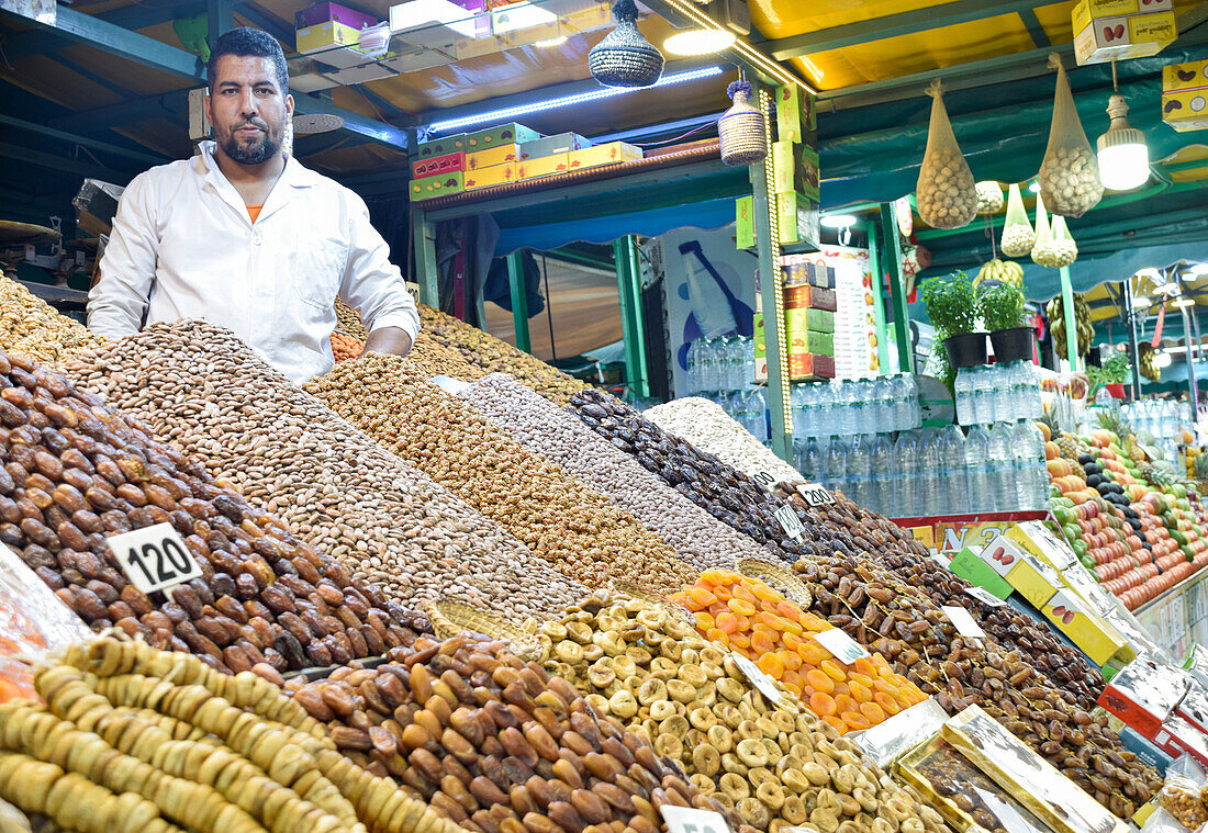 A vendor, appearing Middle-Eastern, looks at the camera while standing behind a colorful display of dates, nuts, and dried fruits at a lively market stall in Marrakesh.