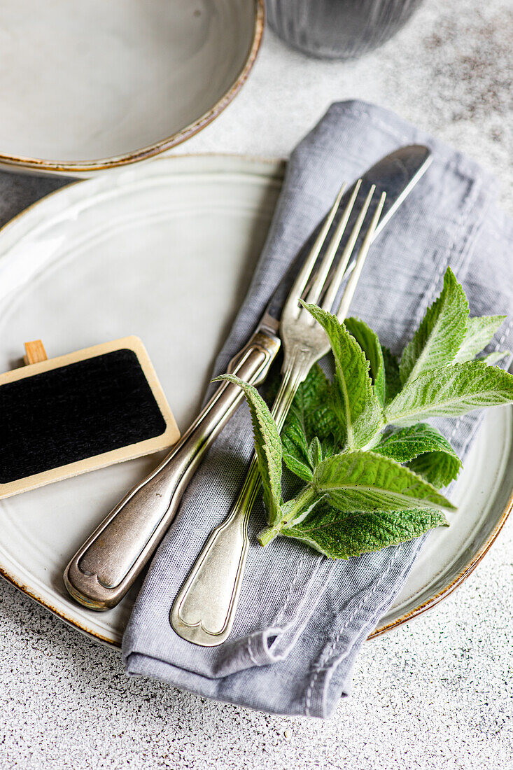 This image captures a beautifully arranged summer table setting centered around a mint theme Fresh mint leaves adorn the scene, complemented by vintage silverware, a rustic name tag, and ceramic plates