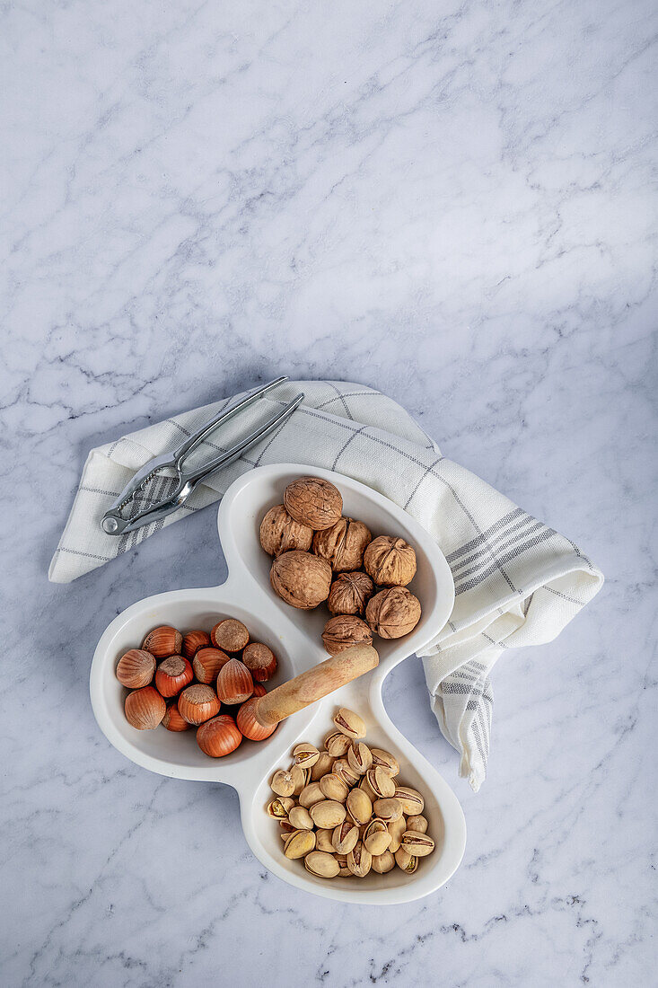 Top view of a variety of whole nuts including walnuts, hazelnuts, and pistachios elegantly presented in a segmented serving dish on a marble surface, accompanied by nutcracker tools.