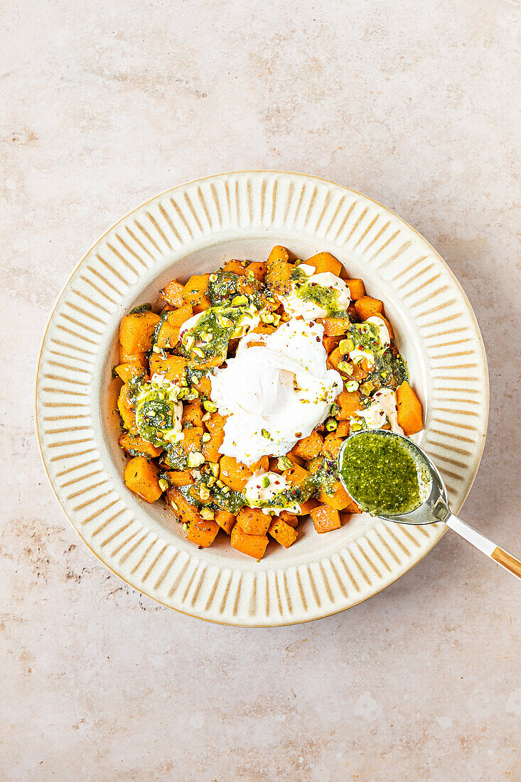 A colorful top view image of roasted butternut squash topped with dollops of creamy mayonnaise and vibrant green pesto, served in a ceramic bowl.