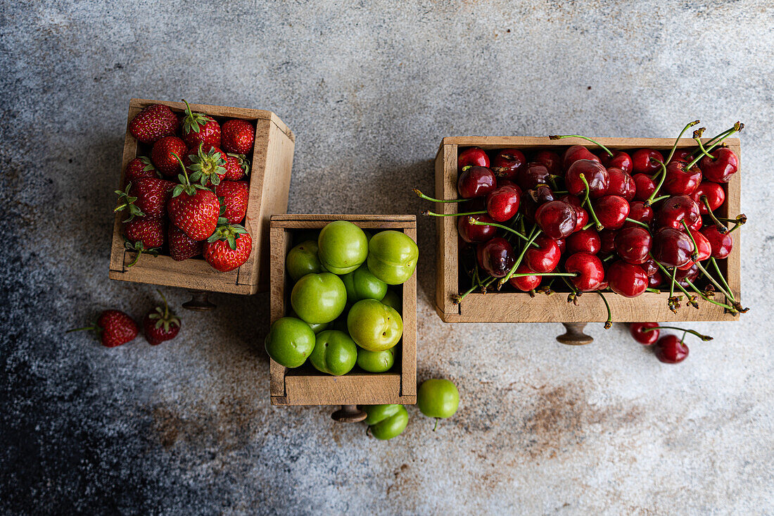 Drei alte Holzkisten präsentieren eine Auswahl frischer Bio-Beeren - grüne Pflaumen, Süßkirschen und Erdbeeren - auf einer rustikalen grauen Oberfläche