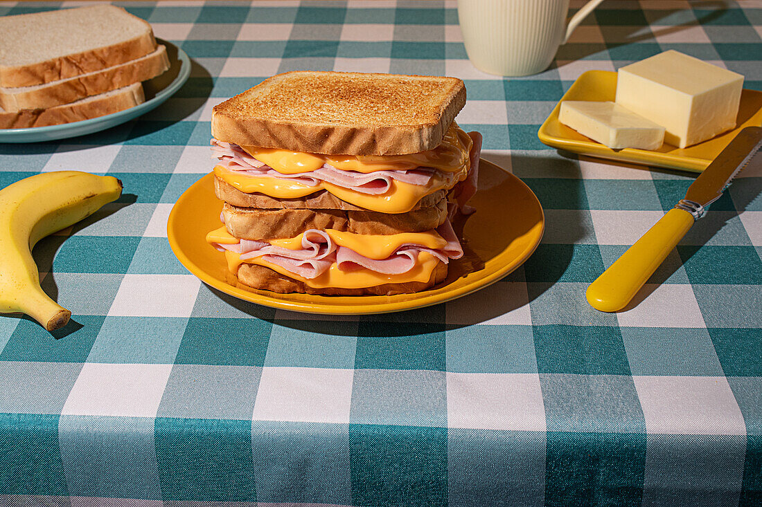 A ham and cheese sandwich on a plate with a banana, butter, and a cup on a checkered blue tablecloth