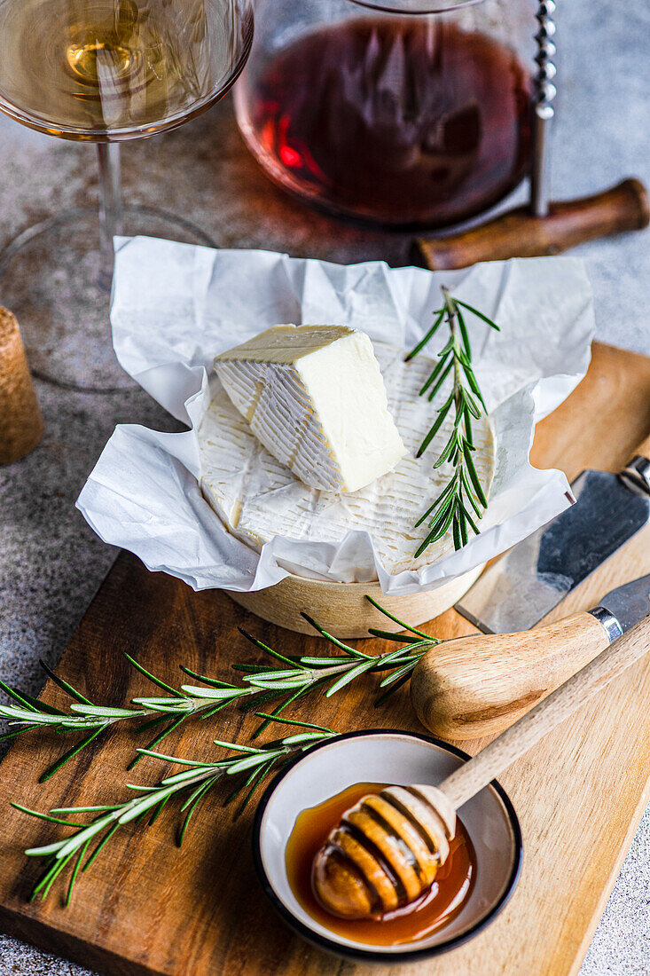 A wedge of Brie cheese on a wooden board, accented with rosemary, next to honey and a glass of white wine.