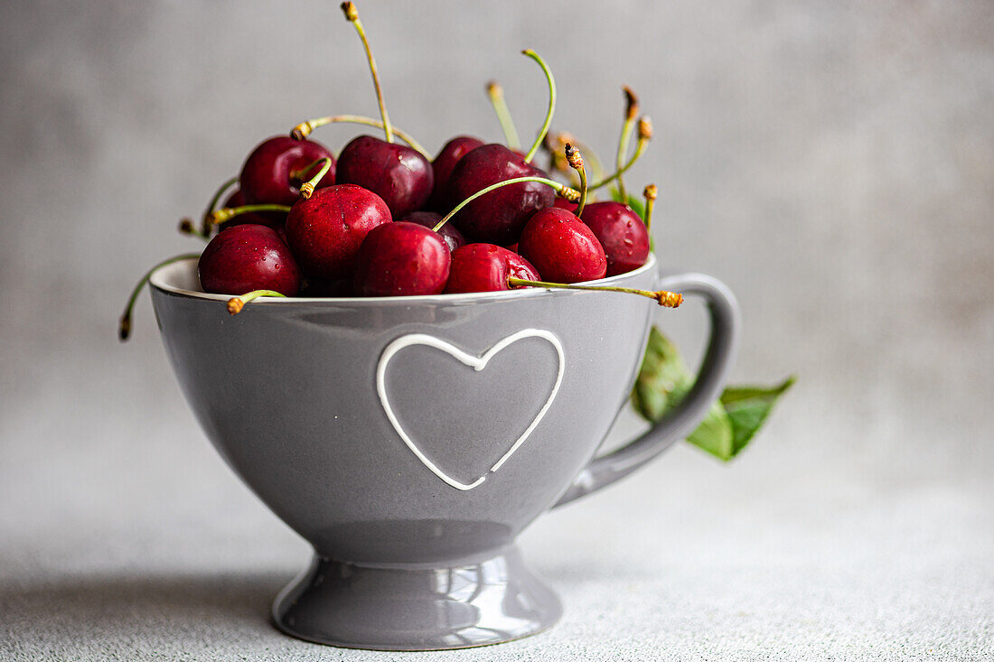 Ripe red cherries in a modern grey bowl with a heart-shaped cutout, placed on a textured grey background. The image captures the vibrant color and freshness of the cherries, suitable for culinary and lifestyle themes.