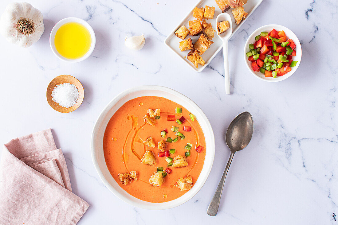 Top view of a traditional Spanish Tomato Gazpacho Soup served with croutons and diced vegetables alongside olive oil and garlic, arranged on a marble surface.