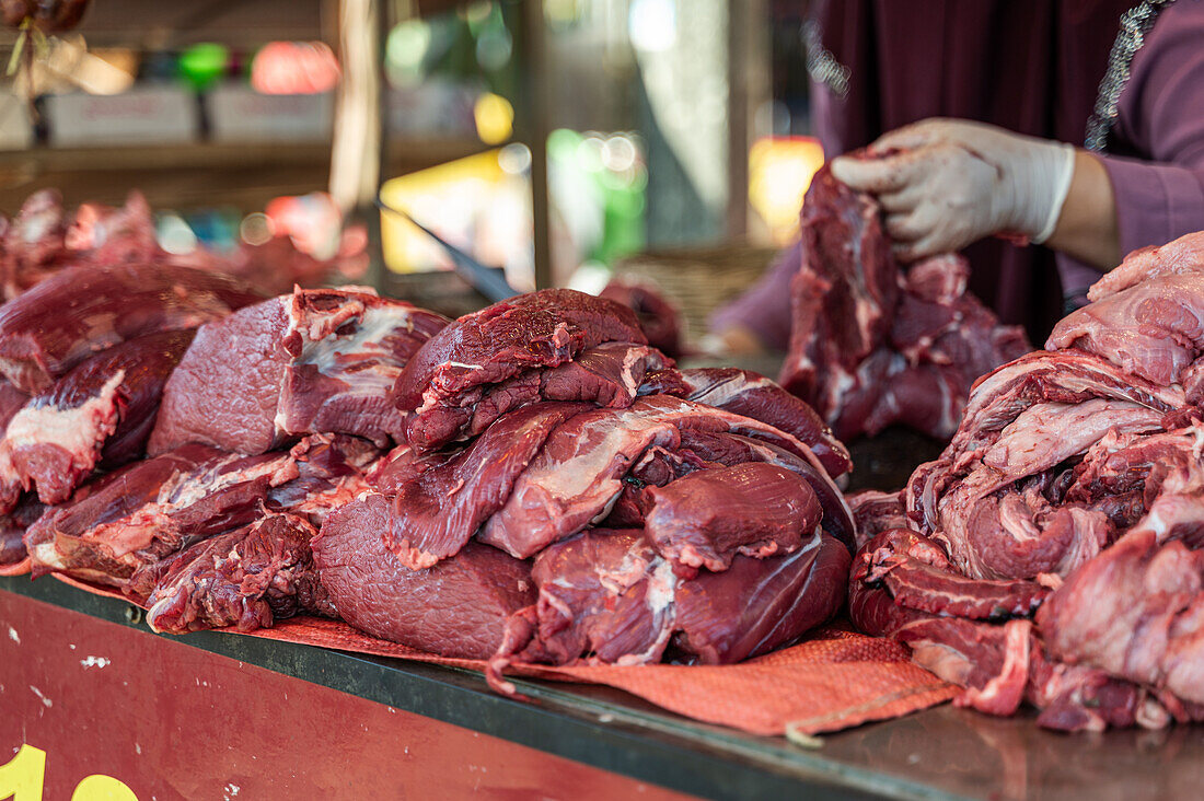 Cropped unrecognizable extensive display of fresh beef cuts at a Bangkok market stall, with a vendor in purple arranging the meat pieces, surrounded by various market paraphernalia.