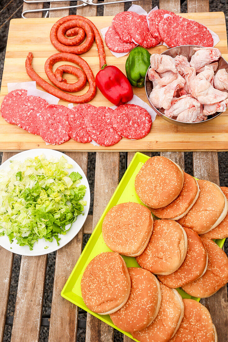 Draufsicht auf eine bunte Vielfalt an rohem Grillgut, darunter Würstchen, Burger-Patties, Hähnchenstücke, rote und grüne Paprika und gehackter Salat neben Sesambrötchen, bereit zum Grillen bei einem Picknick.