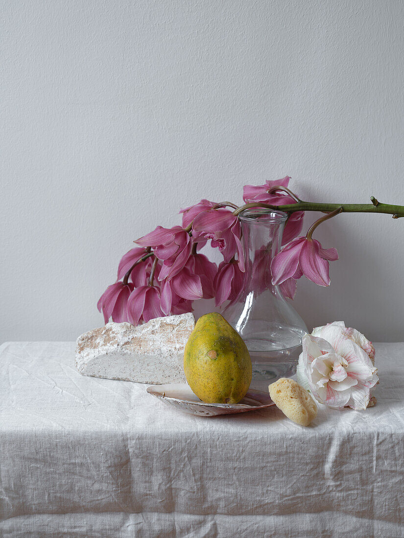 This still life captures the serene atmosphere of a kitchen scene featuring a rustic loaf of bread, a ripe pear, a glass vase filled with pink orchids, and delicate pink flowers on a textured linen tablecloth