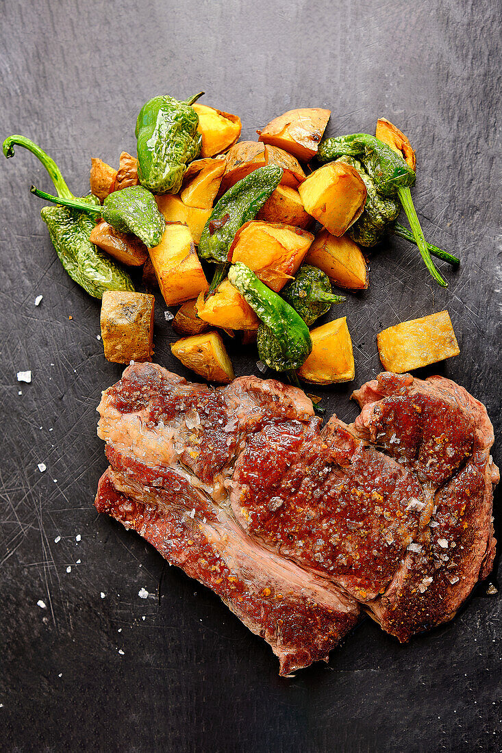 Juicy, cooked steak with coarse salt paired with golden roasted sweet potatoes and blistered green peppers on a dark slate surface.