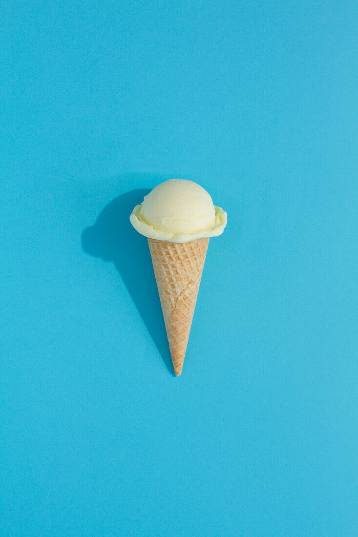 Eine leuchtende cremefarbene Eiskugel sitzt auf einer klassischen Waffeltüte vor einem blauen Hintergrund und wirft einen verspielten Schatten