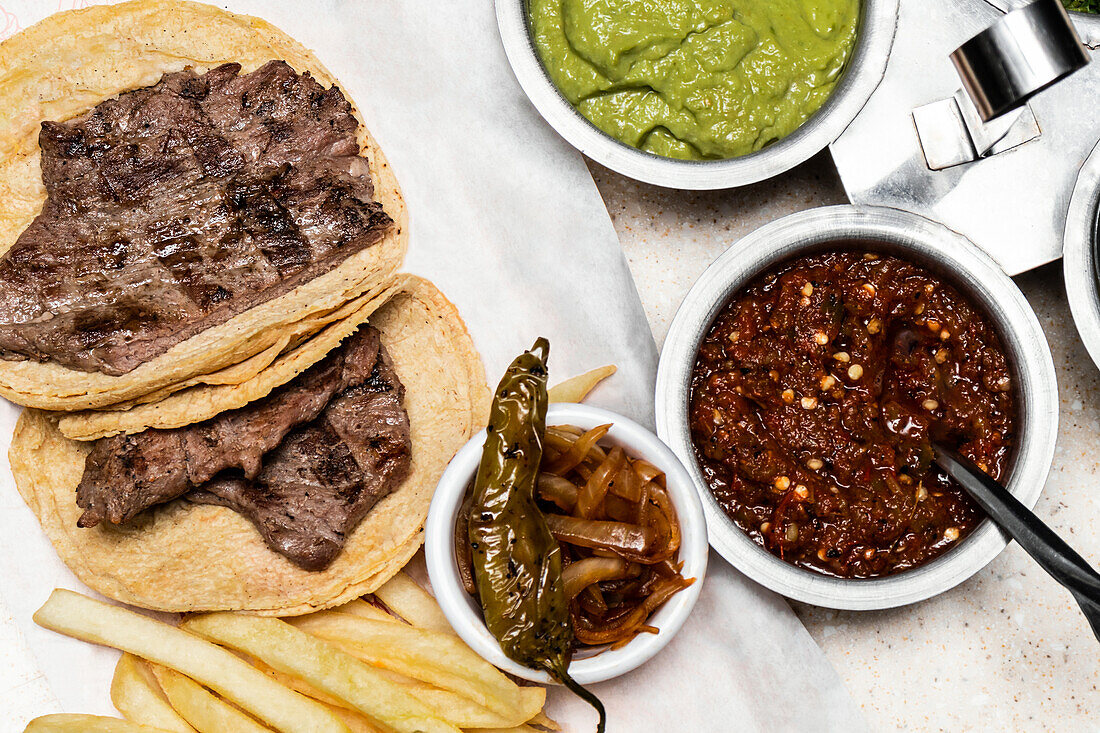 A top view of a traditional Mexican homemade meal featuring grilled steak on corn tortillas, accompanied by guacamole, salsa roja, caramelized onions, and a side of fries.