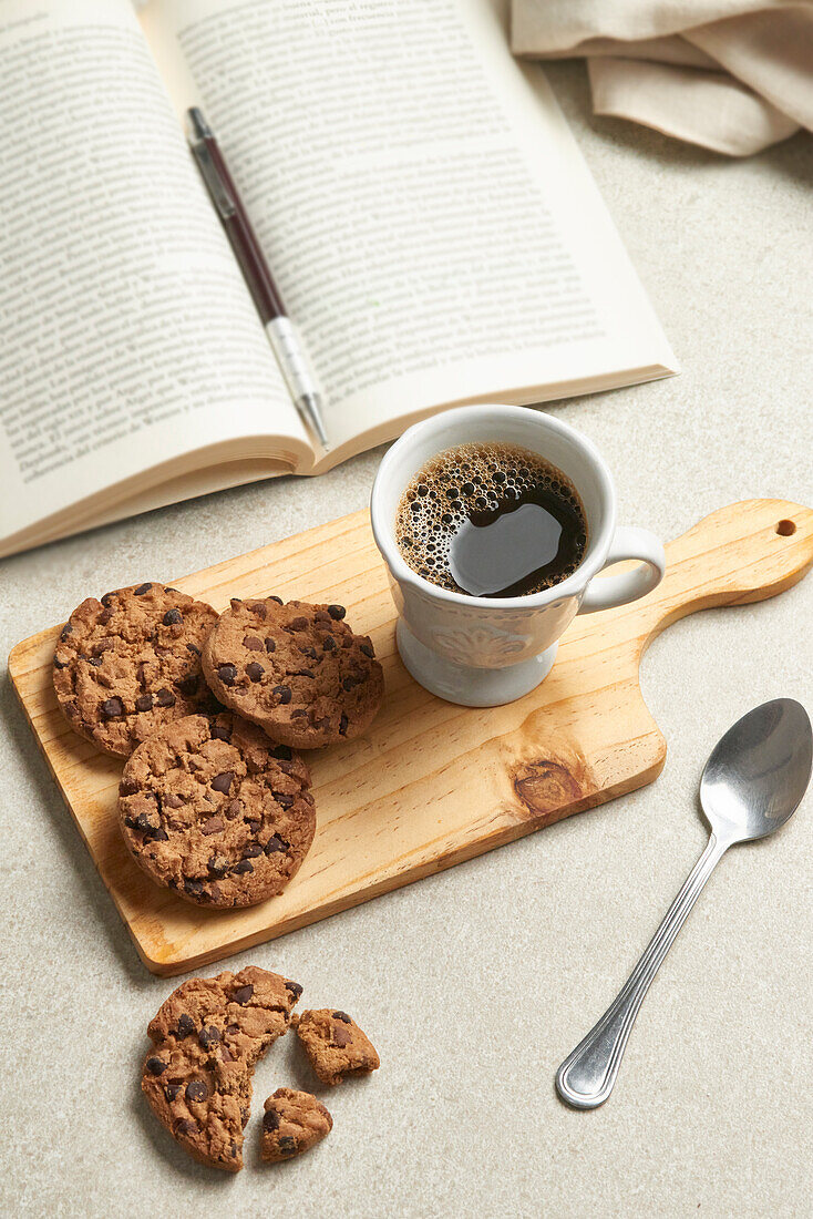 Eine einladende Szene mit einer Tasse Kaffee, einem Stapel Schokoladenkekse auf einem Holzbrett und einem aufgeschlagenen Buch mit einem Stift für eine entspannte Lesezeit