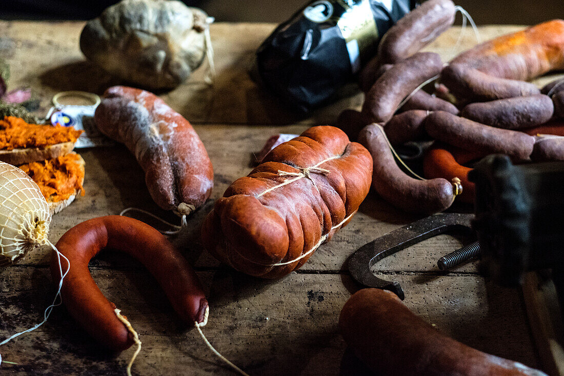 Eine Auswahl an frischen und gepökelten Würsten, darunter die leuchtend rote Sobrasada, die auf einem abgenutzten Holztisch präsentiert wird, begleitet von rustikalem Kochwerkzeug und Gewürzen.