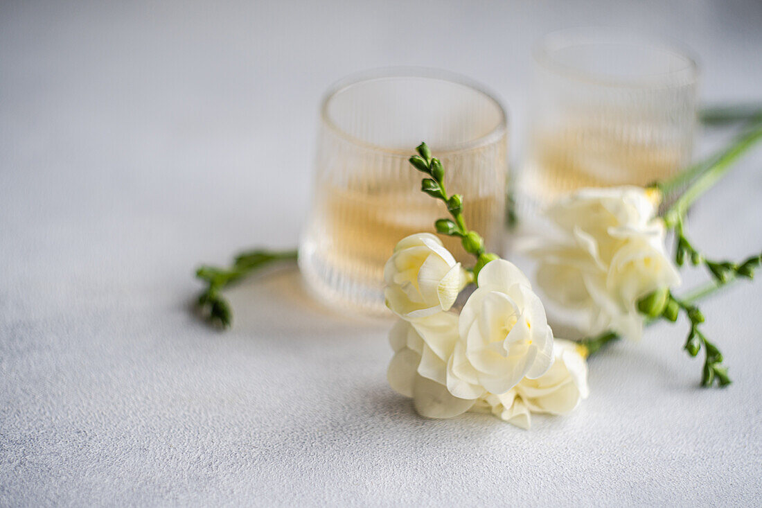 Elegant image showcasing two glasses of peach vodka cocktails accompanied by beautifully arranged white Freesia flowers on a subtle textured background.