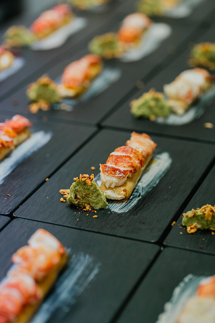 Luxuriöse Hummer-Canapes mit einem Klecks grüner Soße, sorgfältig platziert auf einer schicken schwarzen Schiefertafel für eine Veranstaltung mit Catering