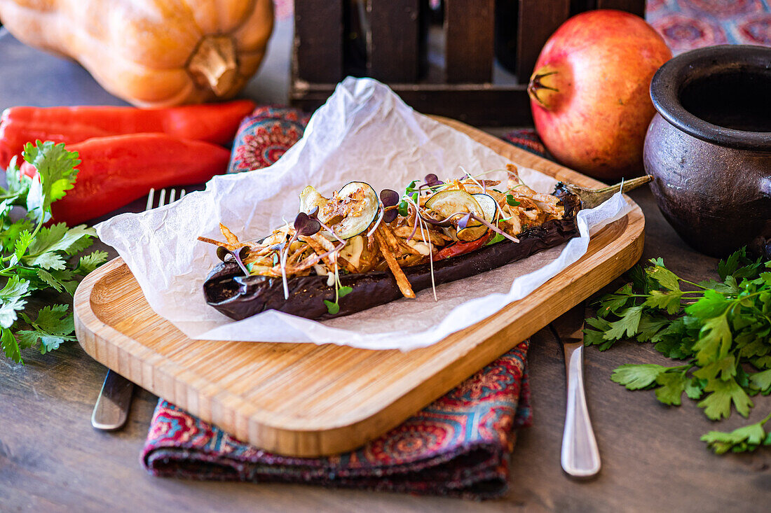 From above, an appetizing stuffed eggplant with cherry tomatoes, bell pepper, onion, garlic, sliced potato, and cheese, beautifully presented on a wooden cutting board, adorned with microgreens and served alongside fresh herbs on rustic tableware.