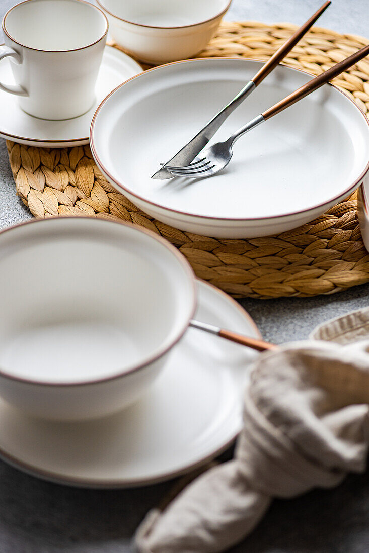 Raffiniertes Tafelservice mit hellen Keramiktellern, elegantem Besteck und einer Tasse auf einem gewebten Tischset.