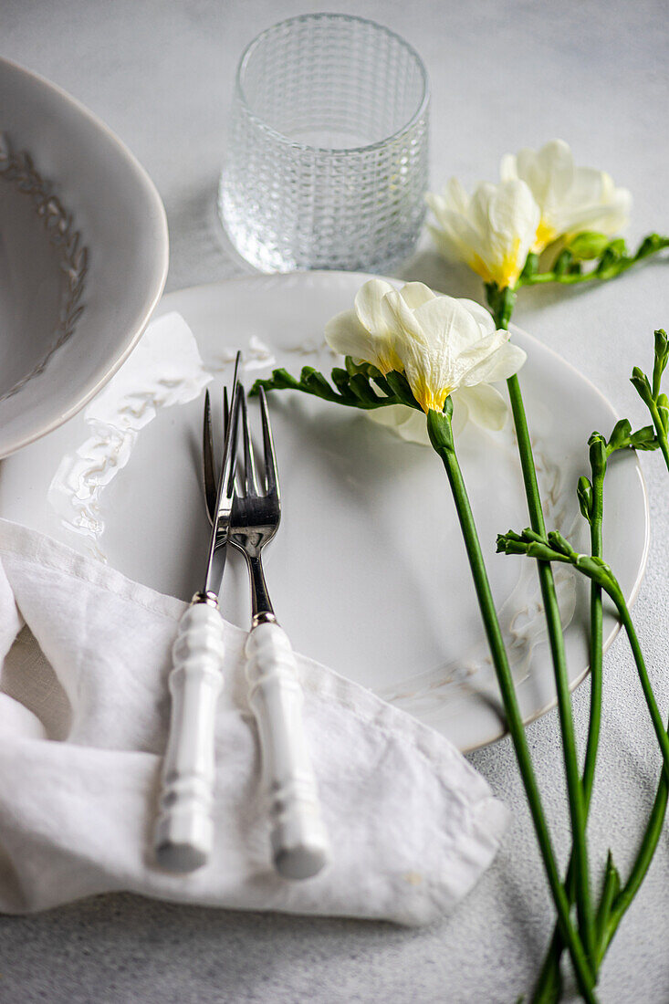 An elegant table with white porcelain dinnerware, polished silverware wrapped in a linen napkin, a textured glass and delicate freesia flowers enhance the refined atmosphere