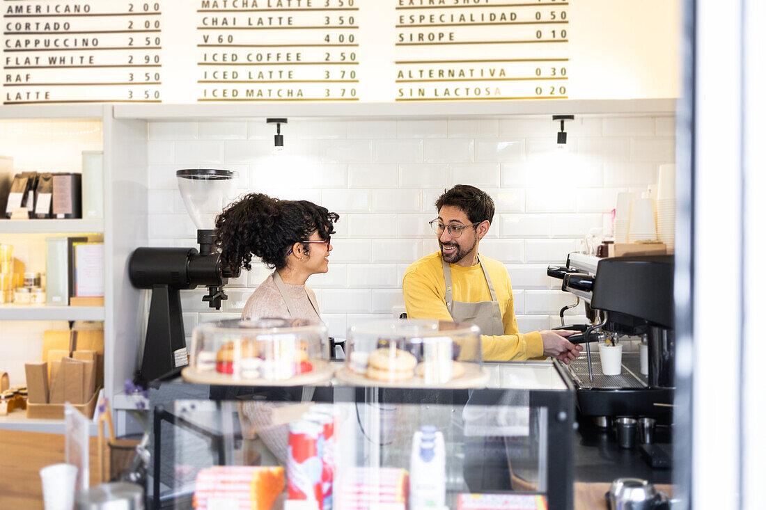 A barista in a yellow apron converses with a customer at a well-lit coffee shop with a visible menu board in the background.