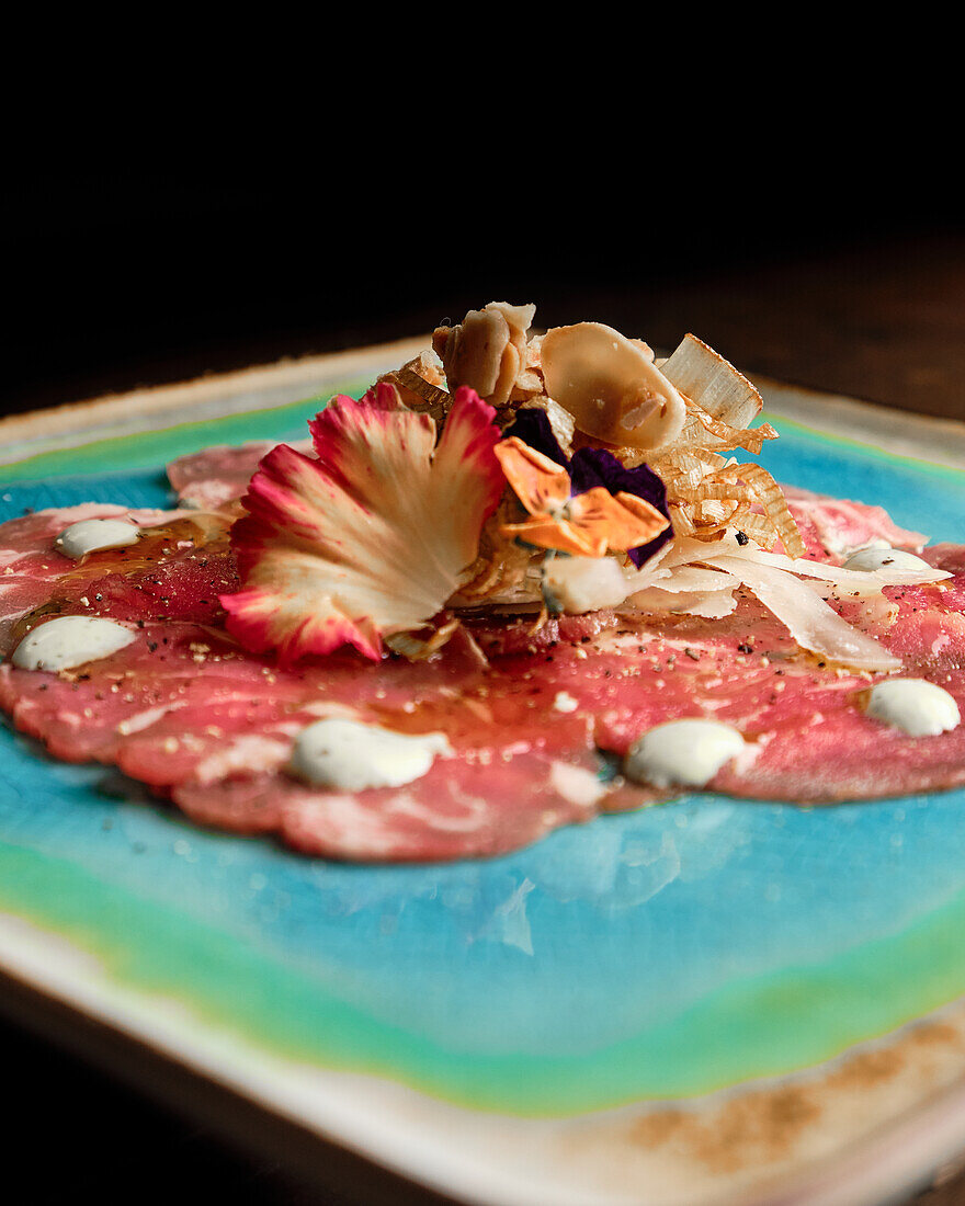 Elegante Präsentation eines Rindercarpaccio, verziert mit essbaren Blumen, Pilzen und Soßentupfern auf einem lebendigen Keramikteller - ideal für Feinschmeckerkonzepte