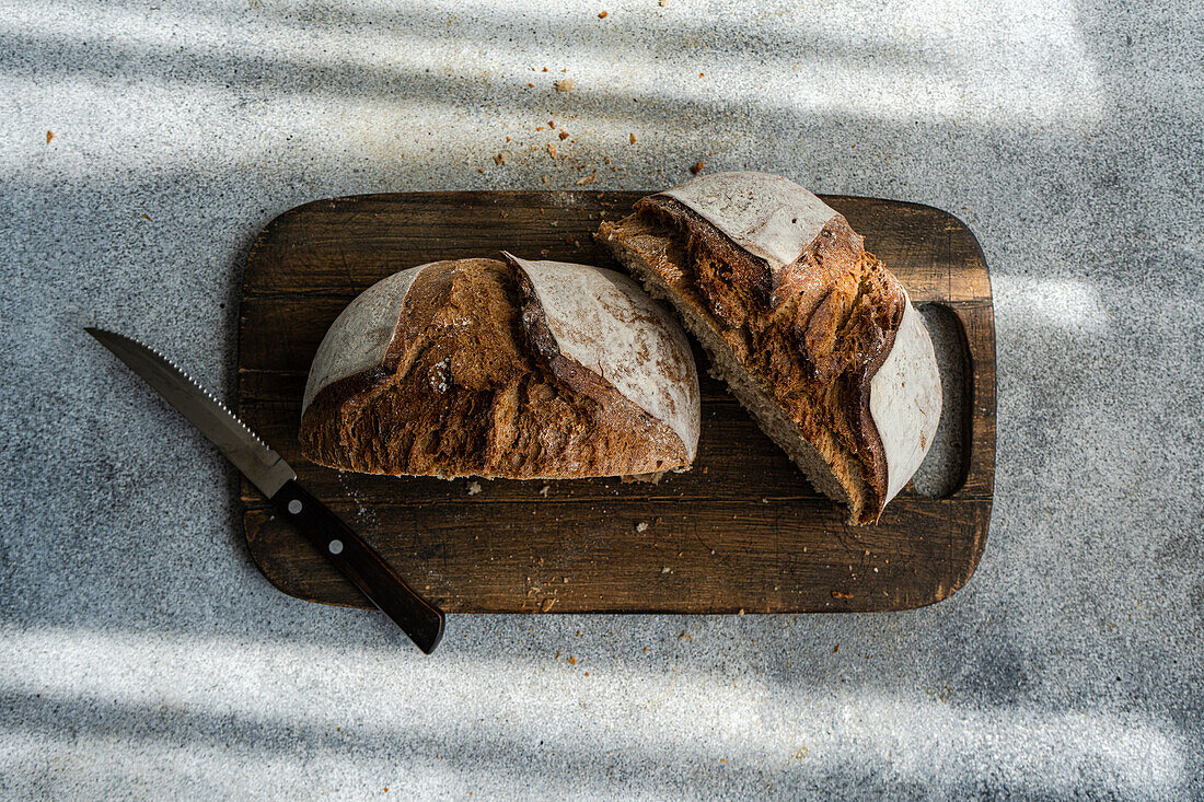 Aufgeschnittenes gesundes Roggensauerteigbrot auf einem rustikalen Holzbrett neben einem gezackten Messer, wobei das natürliche Licht Schatten wirft