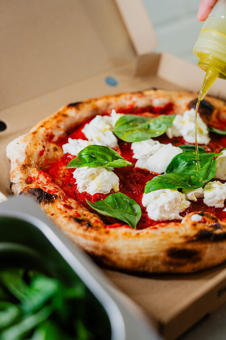 Eine anonyme Person beträufelt eine mit Mozzarella und Basilikum belegte Pizza Margherita mit Olivenöl, die frisch gebacken und in einem Karton zum Mitnehmen serviert wird