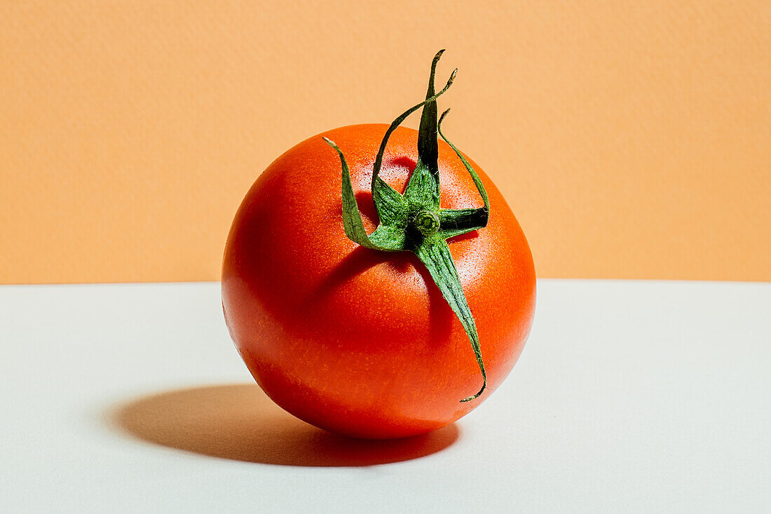 A vibrant red tomato with a fresh green stem is artistically placed against a two-tone orange and white background.