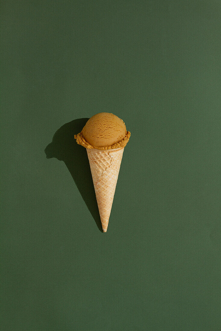 A vibrant coffee ice cream scoop sits atop a classic waffle cone against a green background, casting a playful shadow