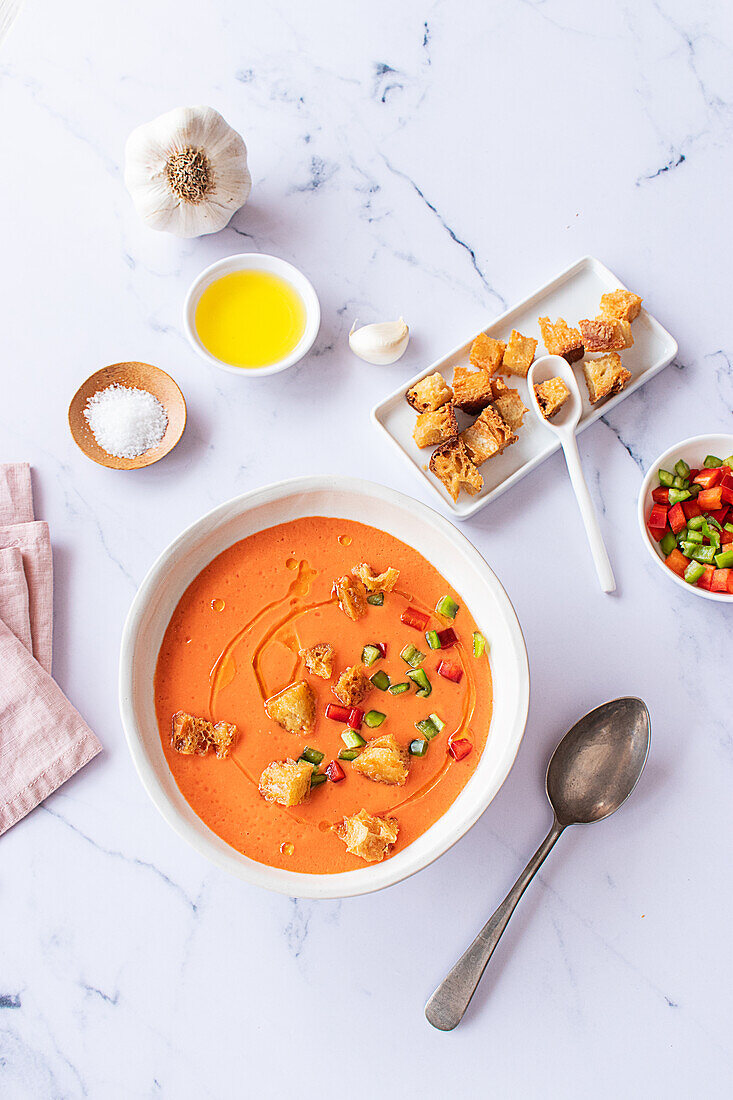 Top view of a traditional Spanish Tomato Gazpacho soup served in a white bowl on a marble background. Garnished with diced vegetables and croutons, accompanied by ingredients like olive oil, garlic, and salt.