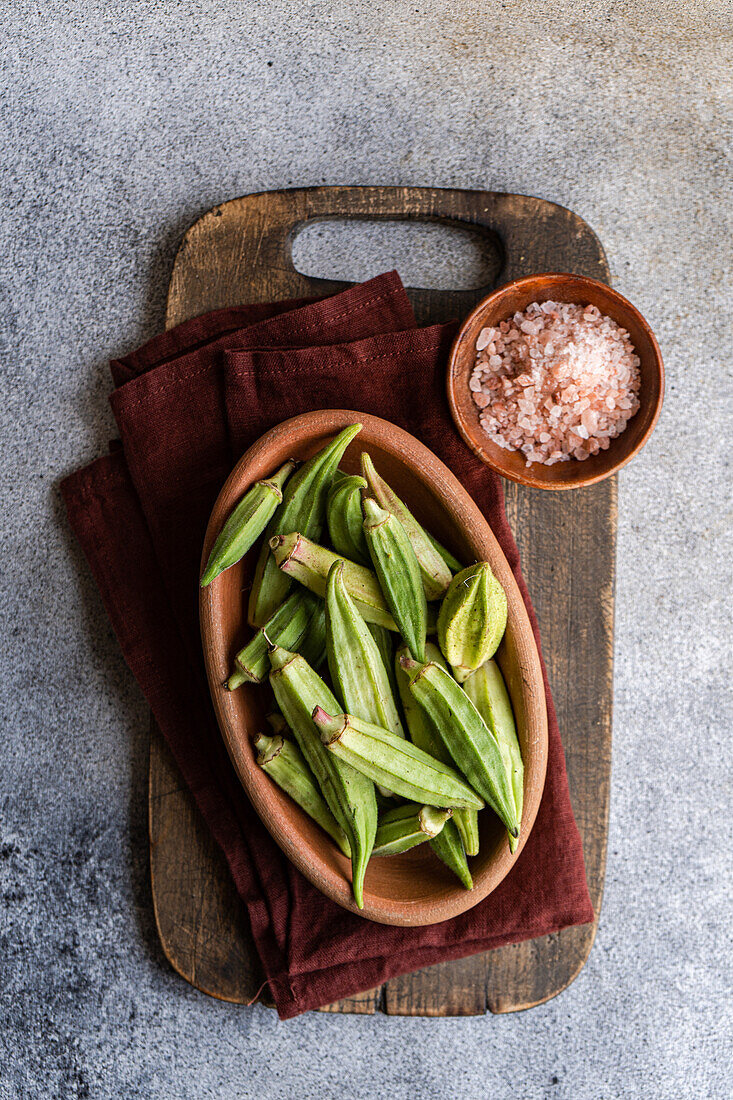 Frische Okra-Bamia-Schoten werden in einer Terrakotta-Schale auf einem hölzernen Schneidebrett vor einem strukturierten grauen Hintergrund präsentiert und verkörpern den rustikalen Charme, der für die mediterrane Küche typisch ist