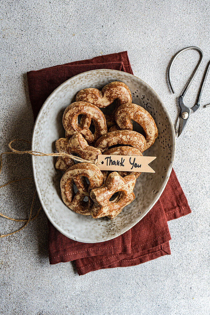 Draufsicht auf selbstgebackene Lebkuchensterne mit Zucker bestäubt, präsentiert in einer Keramikschüssel auf einer strukturierten grauen Oberfläche mit einem Dankeschön-Anhänger, begleitet von einer Schere und einem kastanienbraunen Tuch.