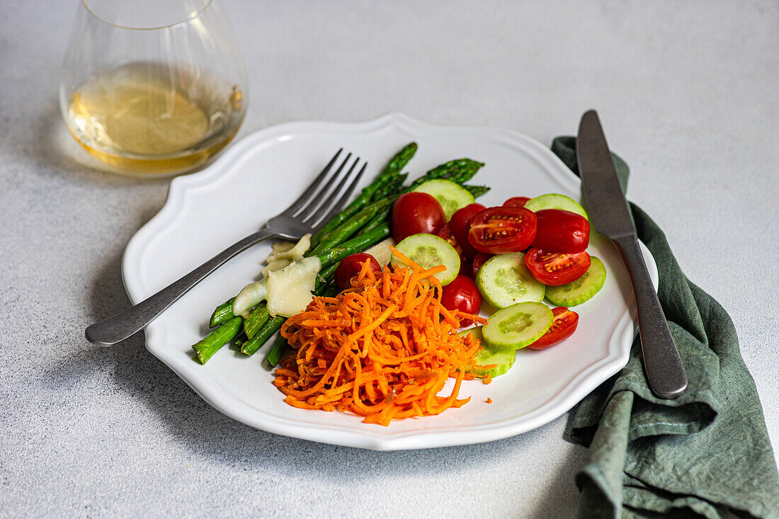 Healthy vegetable salad featuring asparagus with cheese, halved cherry tomatoes, cucumber slices, and grated carrots on a white plate with dressing and cutlery beside.