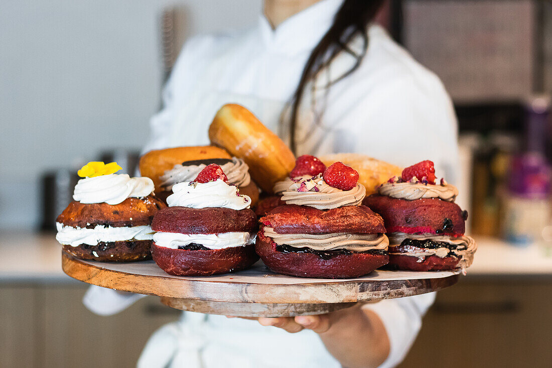 Crop anonymous female baker in uniform standing in bakery with tray of tasty vegan Berliners with whipped cream filling