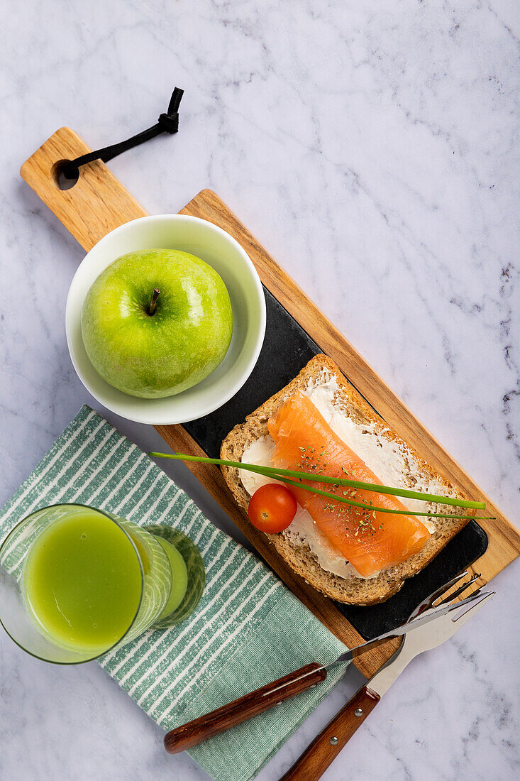 Top view of a healthy food layout featuring a green apple in a bowl, a slice of bread topped with smoked salmon and cream cheese, a cherry tomato, cucumber and apple juice in a glass, and utensils arranged neatly on a marble background.