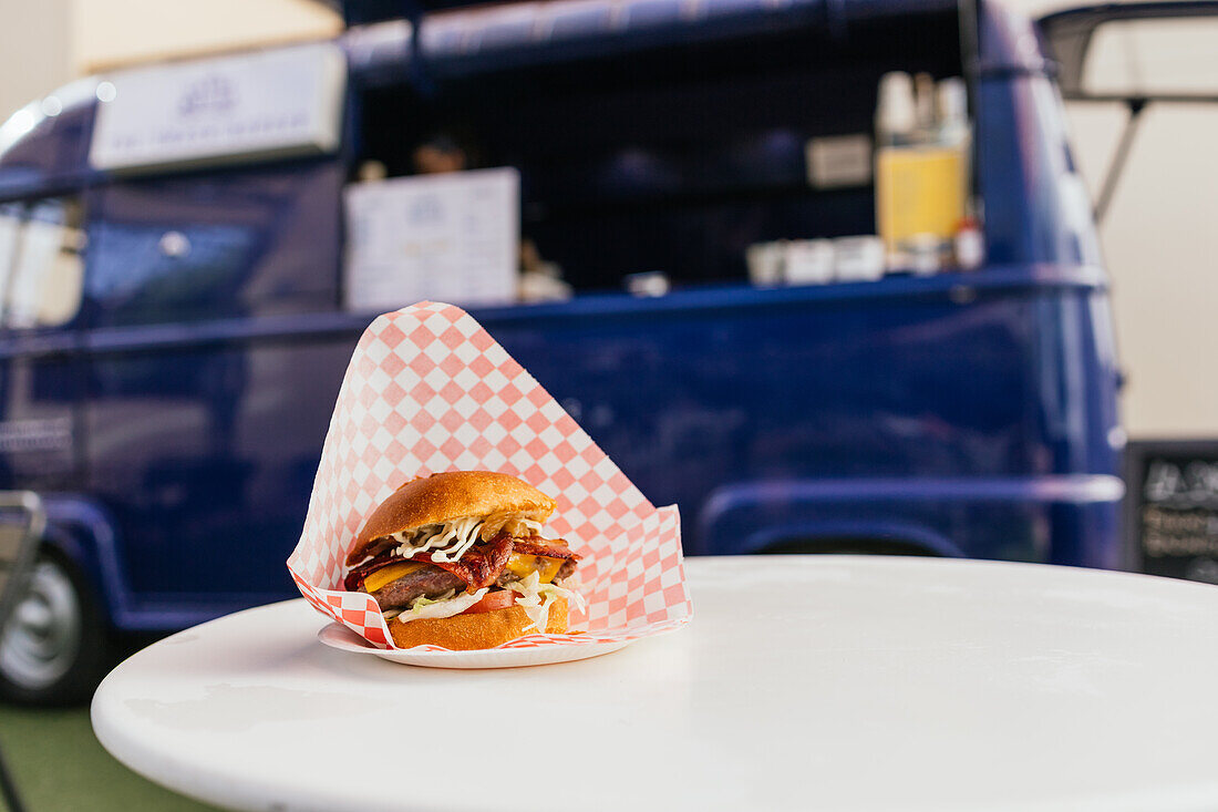 Ein köstlicher Gourmet-Burger, eingewickelt in klassisches kariertes Papier, liegt auf einem weißen Tisch, mit einem blauen Foodtruck im Hintergrund, der eine Street-Food-Szene unterstreicht