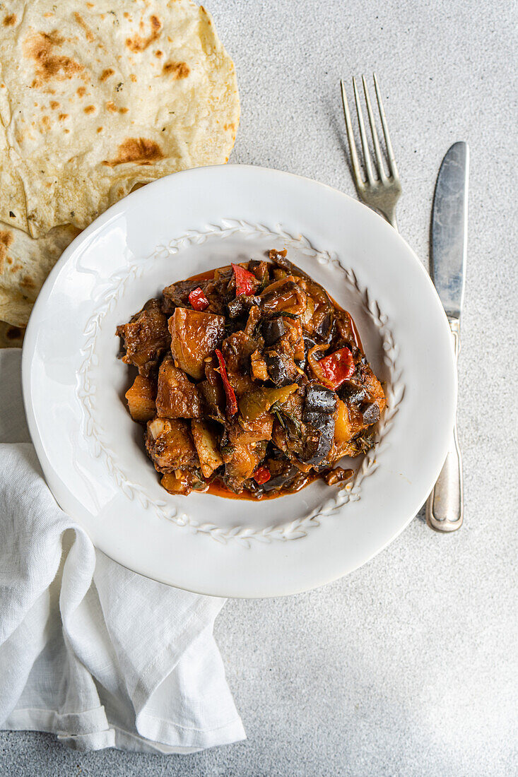 Ein deftiger Gemüseeintopf mit Auberginen, Tomaten, Kartoffeln, Paprika, Zwiebeln, Knoblauch und Basilikum, gepaart mit Fladenbrot auf einem weißen Teller, perfekt für eine nahrhafte Mahlzeit