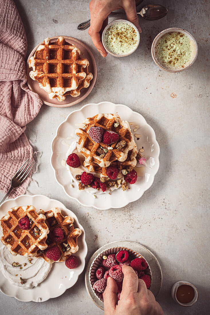 A cozy baking scene featuring waffles topped with raspberries and cream, along with cups of matcha latte